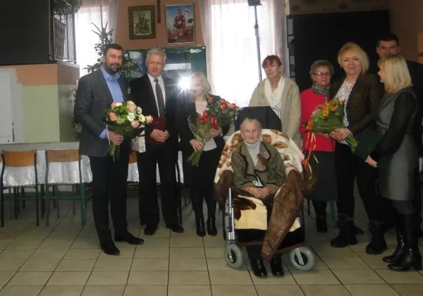 Uroczystość z okazji setnych urodzin mieszkanki powiatu płockiego, na której obecni są bliscy oraz przedstawiciele lokalnych władz, wręczający kwiaty i gratulacje.