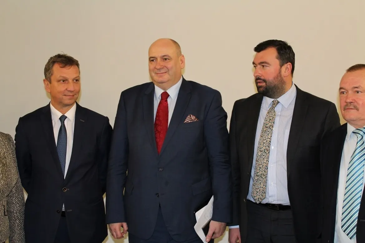 Na zdjęciu grupa mężczyzn w garniturach oraz jedna kobieta, stojących w prostym wnętrzu, dyskutujących na temat podziału województwa mazowieckiego.