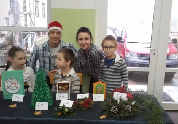 Grupa dzieci i dorosłych pozują przy stoisku z rękodziełem świątecznym na charytatywnym wydarzeniu.