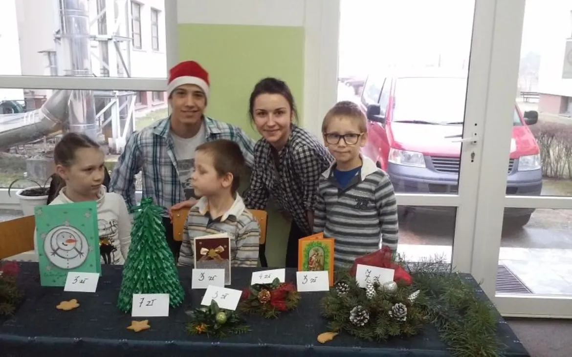 Grupa dzieci i dorosłych pozują przy stoisku z rękodziełem świątecznym na charytatywnym wydarzeniu.