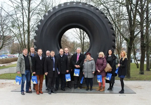 Grupa osób stoi przed dużym pomnikiem w kształcie opony, trzymając torby z logo, w kontekście rozwoju Warmińsko-Mazurskiej Specjalnej Strefy Ekonomicznej w powiecie płockim.