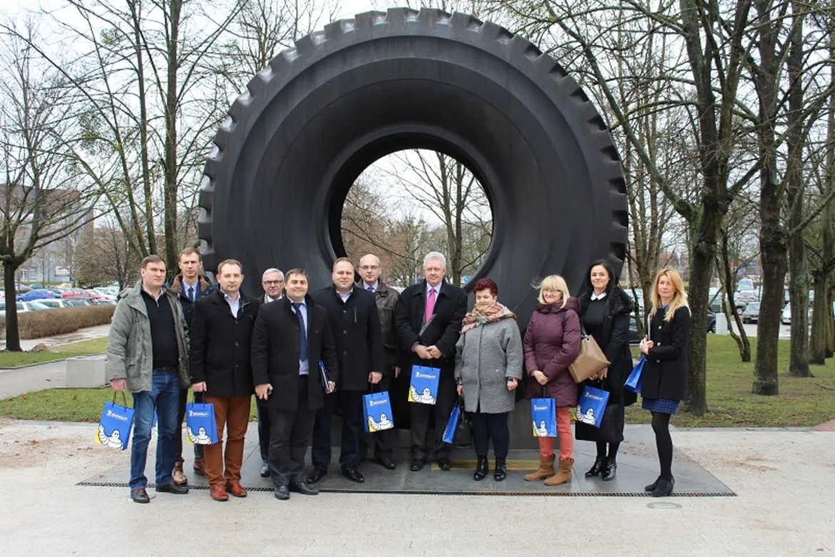 Grupa osób stoi przed dużym pomnikiem w kształcie opony, trzymając torby z logo, w kontekście rozwoju Warmińsko-Mazurskiej Specjalnej Strefy Ekonomicznej w powiecie płockim.