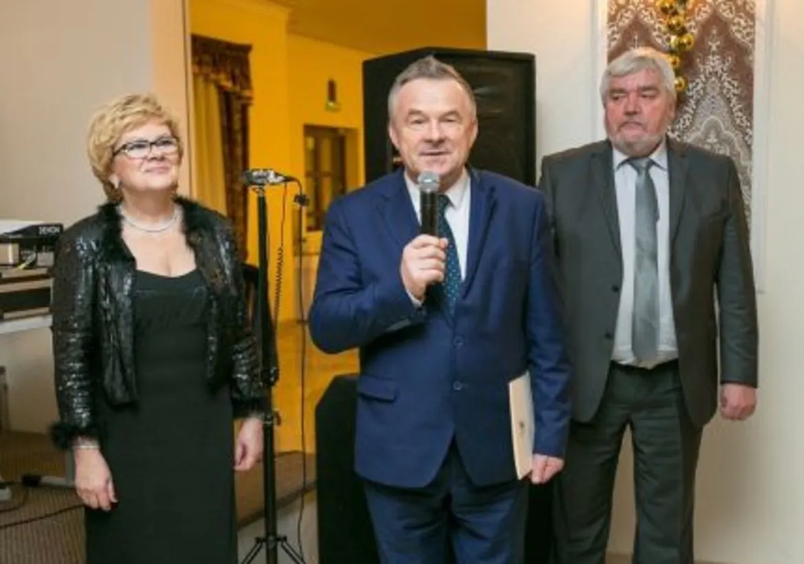 Na zdjęciu widać trzech dorosłych: kobietę w czarnej sukni, która stoi z boku, mężczyznę wygłaszającego przemówienie z mikrofonem oraz drugiego mężczyznę w szarym garniturze, obserwującego sytuację.