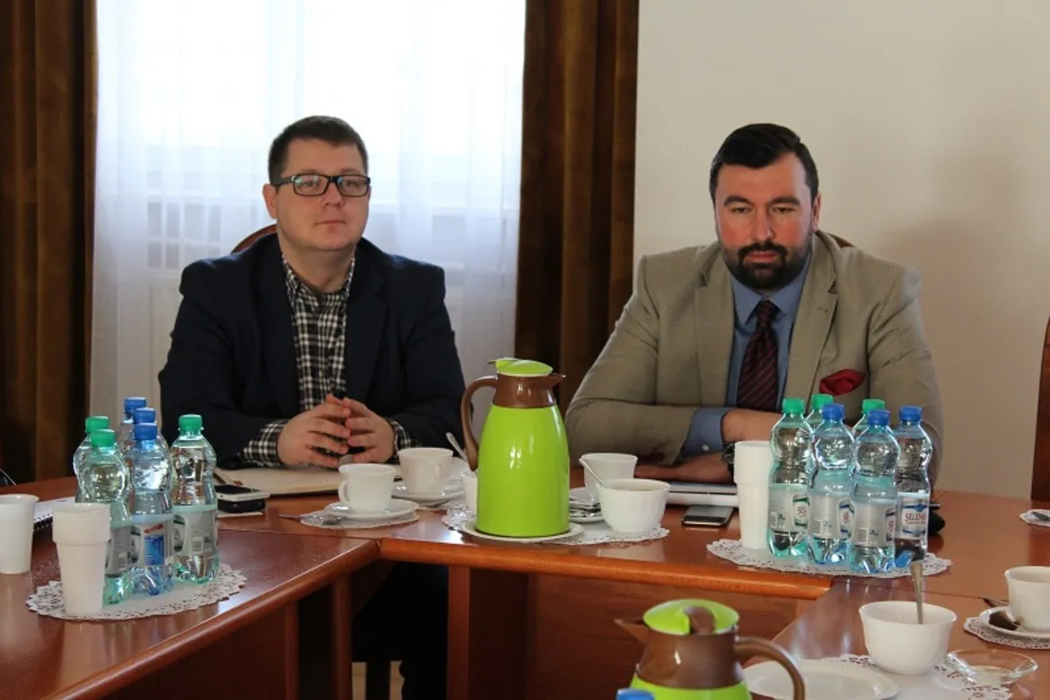 Na zdjęciu dwóch mężczyzn siedzi przy stole w trakcie spotkania dotyczącego przebudowy drogi powiatowej nr 2974W Soczewka – Łąck, z filiżankami i butelkami wody na stole.