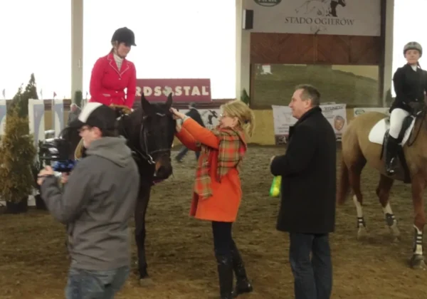 Na zdjęciu widać zawodniczkę w czerwonym stroju, siedzącą na koniu, podczas przygotowań do Halowych Zawodów w Skokach przez Przeszkody w Stadzie Ogierów w Łącku, z osobą pomagającą jej w stroju oraz innymi uczestnikami w tle.
