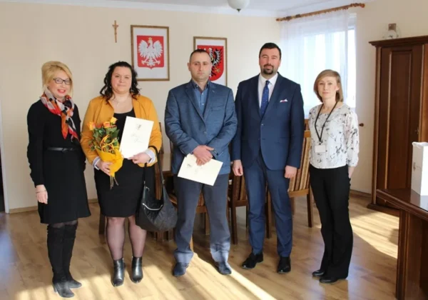 Pięć osób (trzy kobiety i dwóch mężczyzn) w formalnych ubraniach stoi w gabinecie Starosty; jedna kobieta trzyma bukiet kwiatów i kartkę, a w tle widać krzyż i herby państwowe.