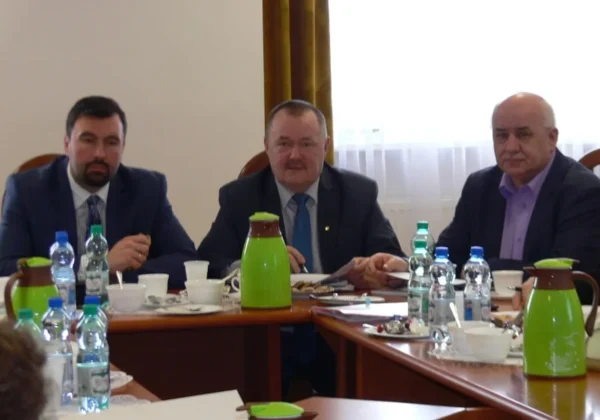 Trzech mężczyzn w garniturach siedzi przy stole w sali konferencyjnej podczas spotkania Zarządu Powiatu w Płocku, na stole widoczne są butelki wody, filiżanki i zielone dzbanki.