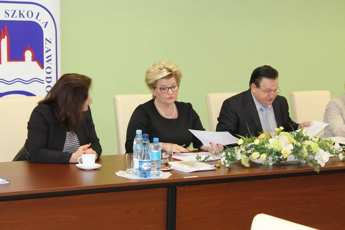 Na zdjęciu przedstawiono uczestników nadzwyczajnego posiedzenia Konwentu Państwowej Wyższej Szkoły Zawodowej w Płocku, siedzących przy stole z dokumentami i kwiatami.