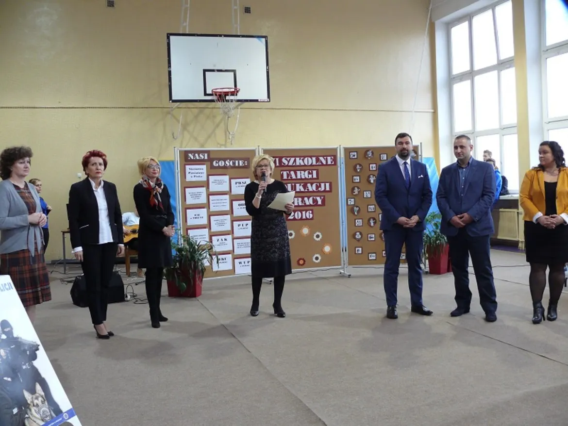 VI Szkolne Targi Edukacji i Pracy w Zespole Szkół im. Stanisława Staszica w Gąbinie, z prezentacją mówców stojących przy tablicy informacyjnej.