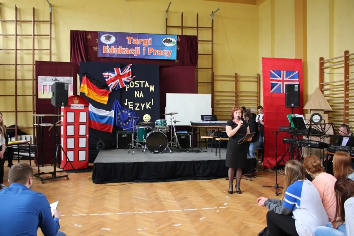 Na scenie targów edukacyjnych „Od edukacji do pracy” odbywa się prezentacja, podczas której mówca stoi przed zebranym audytorium, a w tle wiszą flagi różnych krajów.