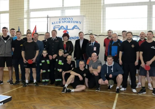 Uczestnicy I Turnieju Tenisa Stołowego Strażaków zebrani w sali sportowej, z flagami i banerem Gminnego Klubu Sportowego Fala.