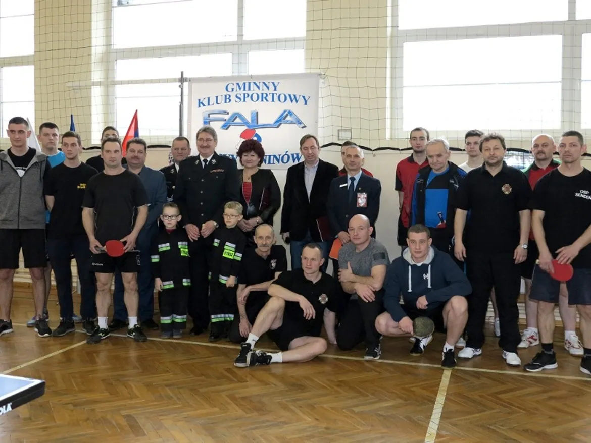 Uczestnicy I Turnieju Tenisa Stołowego Strażaków zebrani w sali sportowej, z flagami i banerem Gminnego Klubu Sportowego Fala.