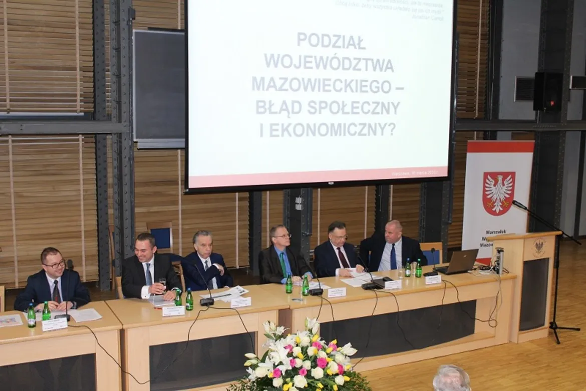 Na zdjęciu przedstawiono panel dyskusyjny dotyczący podziału Województwa Mazowieckiego, z prominentnymi uczestnikami oraz prezentacją na temat społecznych i ekonomicznych skutków tego podziału.