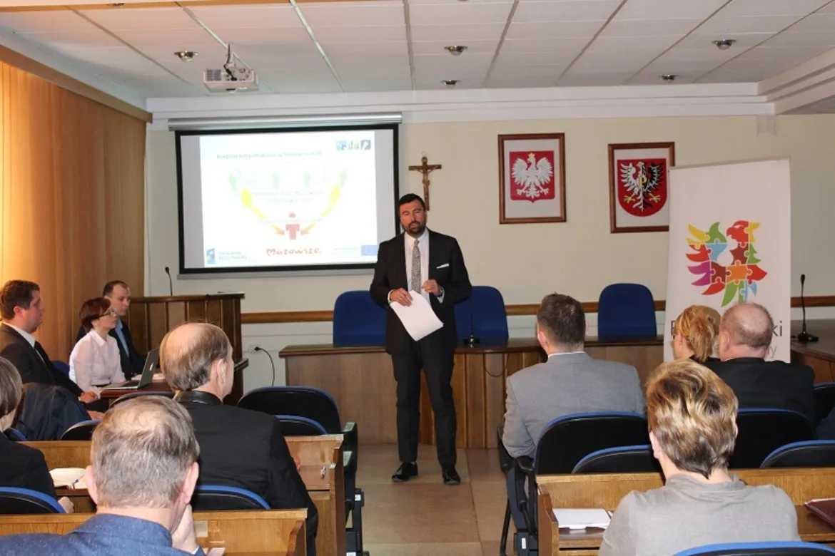 Na zdjęciu widać wystąpienie mężczyzny w garniturze podczas konferencji podsumowującej projekt „INTERNET DLA MAZOWSZA”, z uczestnikami siedzami na widowni.