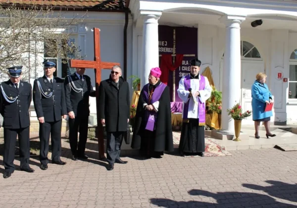 Uczestnicy Drogi Krzyżowej w Goślicach stoją w grupie przed kościołem, trzymając krzyż oraz biorąc udział w uroczystości religijnej.