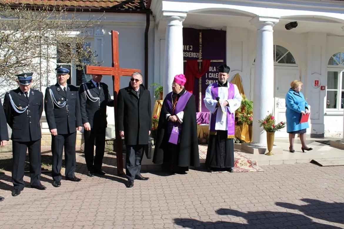 Uczestnicy Drogi Krzyżowej w Goślicach stoją w grupie przed kościołem, trzymając krzyż oraz biorąc udział w uroczystości religijnej.