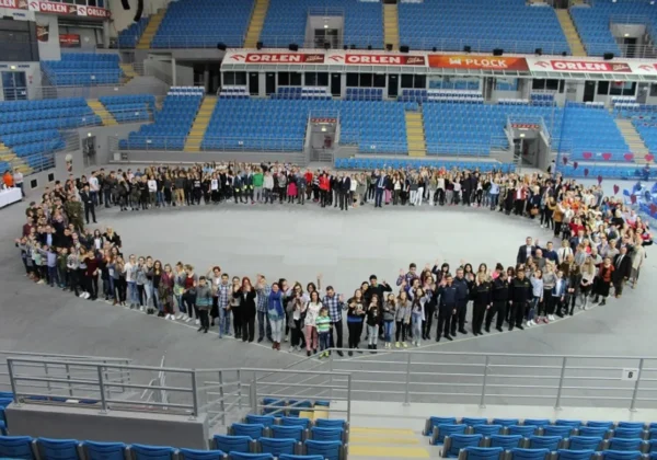 Grupa ludzi w kształcie serca w hali sportowej, z podziękowaniami dla Wielkiej Orkiestry Świątecznej Pomocy.