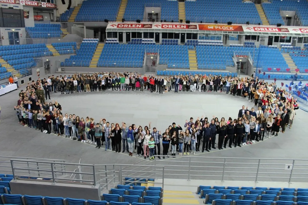 Grupa ludzi w kształcie serca w hali sportowej, z podziękowaniami dla Wielkiej Orkiestry Świątecznej Pomocy.