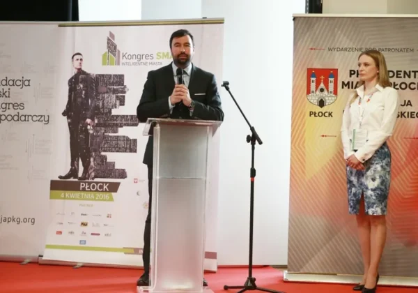 Na zdjęciu przedstawiono mężczyznę przemawiającego z mównicy podczas Kongresu SMART – Inteligentne Miasta w Płocku, z towarzyszącą mu kobietą i banerami informacyjnymi w tle.