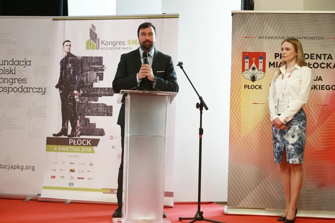 Na zdjęciu przedstawiono mężczyznę przemawiającego z mównicy podczas Kongresu SMART – Inteligentne Miasta w Płocku, z towarzyszącą mu kobietą i banerami informacyjnymi w tle.