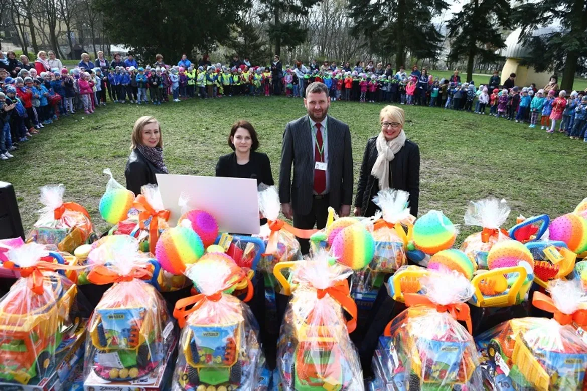 Na zdjęciu widoczna jest grupa dzieci i dorosłych zgromadzonych na finale Konkursu na Najpiękniejszą Marzannę, z wyeksponowanymi kolorowymi nagrodami na pierwszym planie.