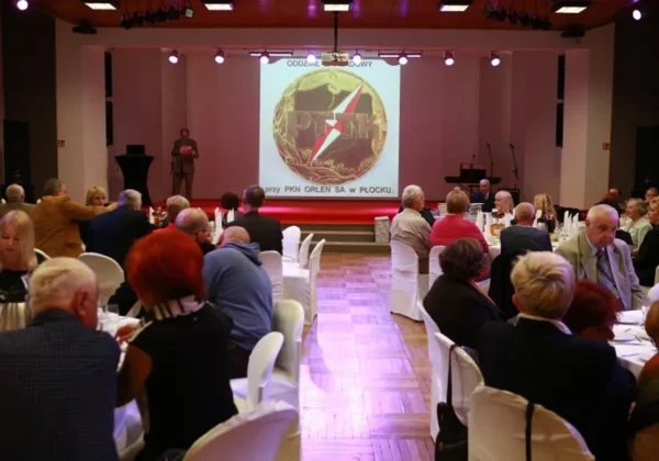 Uroczystość z okazji 50-lecia Oddziału Zakładowego PTTK przy PKN ORLEN, gdzie goście siedzą przy okrągłych stołach, a na scenie prowadzący wygłasza przemówienie przy dużym ekranie z logiem PTTK.