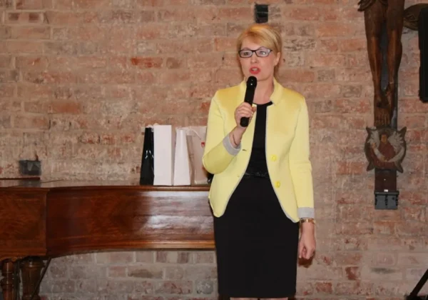 Kobieta w żółtym żakiecie trzyma mikrofon i przemawia na scenie przy ceglanej ścianie, z fortepianem i prezentami w tle.