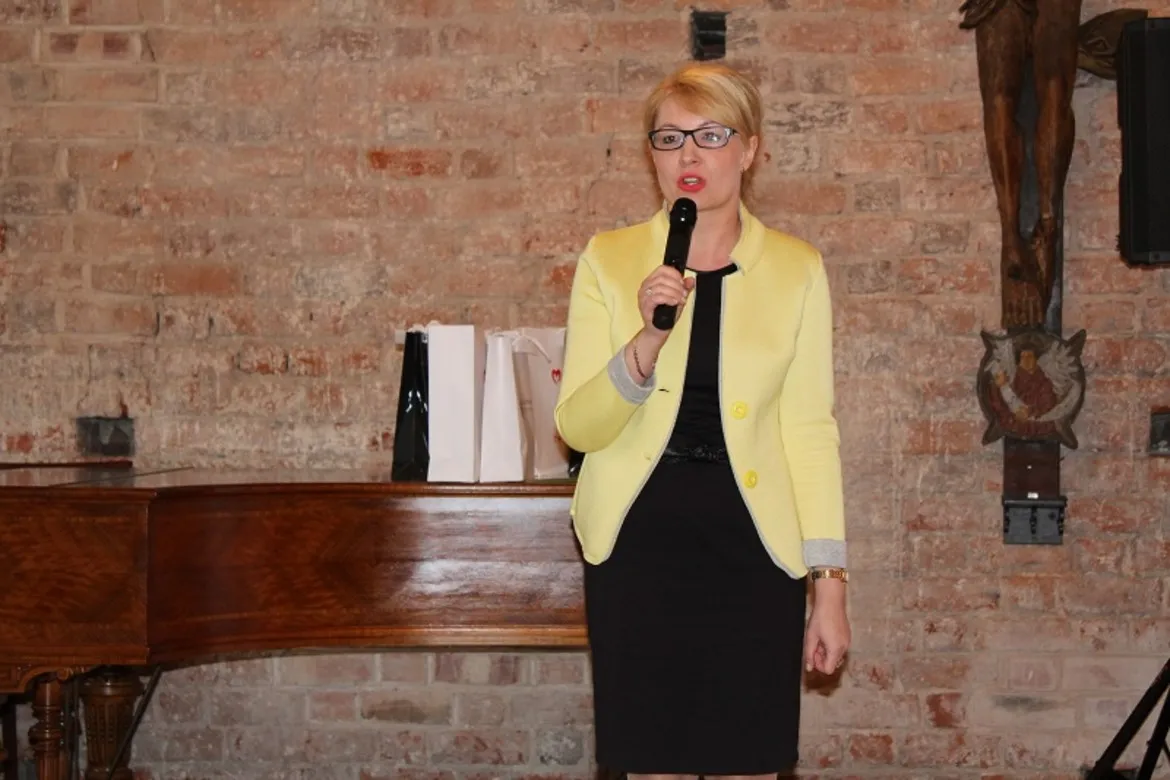 Kobieta w żółtym żakiecie trzyma mikrofon i przemawia na scenie przy ceglanej ścianie, z fortepianem i prezentami w tle.