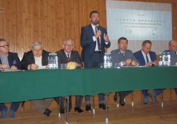 Panel dyskusyjny podczas debaty o bezpieczeństwie publicznym w Bulkowie: grupa mężczyzn w garniturach siedzi za zielonym obrusem, jeden z nich stoi i przemawia do mikrofonu, na ekranie w tle widoczny jest plakat.