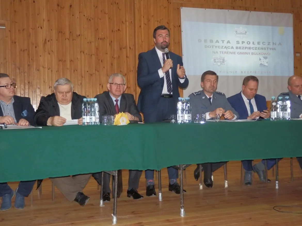 Panel dyskusyjny podczas debaty o bezpieczeństwie publicznym w Bulkowie: grupa mężczyzn w garniturach siedzi za zielonym obrusem, jeden z nich stoi i przemawia do mikrofonu, na ekranie w tle widoczny jest plakat.