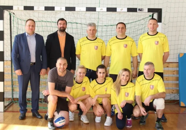 Grupa drużyn mieszanych w żółtych koszulkach z herbem Powiatu Płockiego stoi i klęczy przy boiskowej siatce w hali podczas VI Mazowieckiego Turnieju Piłki Siatkowej Drużyn Mieszanych o Puchar Starosty Powiatu Płockiego.