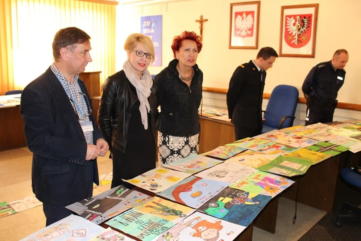 Grupa osób ogląda kolorowe prace plastyczne rozłożone na stole podczas konkursu „W trosce o nasze bezpieczeństwo”.