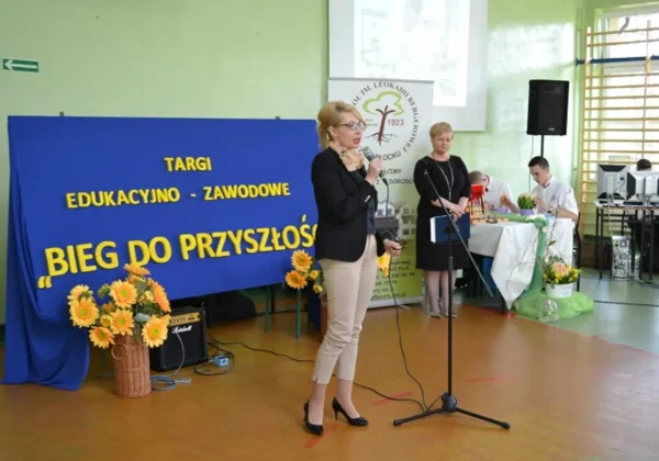 Podczas Targów Edukacyjno-Zawodowych w Zespole Szkół im. Leokadii Bergerowej w Płocku dwie kobiety prowadzą wystąpienie z mikrofonami na tle niebieskiej dekoracji z napisem „TARGI EDUKACYJNO-ZAWODOWE BIEG DO PRZYSZŁOŚCI” i kwiatowych ozdób.