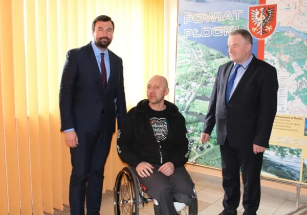 Trzech mężczyzn stoi w pomieszczeniu przy dużej mapie Powiatu Płockiego, a środkowy siedzi na wózku inwalidzkim.