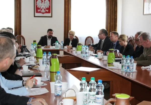 Grupa osób w formalnych strojach siedzi przy długim stole konferencyjnym podczas posiedzenia Komisji Bezpieczeństwa i Porządku, z dokumentami, filiżankami i zielonymi termosami na stole, w sali z godłem Polski na ścianie.