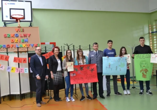 Grupa uczniów i nauczycieli wraz z funkcjonariuszem stoi na sali gimnastycznej podczas VIII Szkolnego Dnia Profilaktyki „Nie dla nałogów – tak dla pasji i działania”, prezentując kolorowe plakaty i banery.