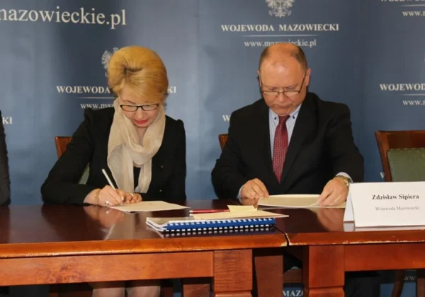 Podczas uroczystego podpisywania umów na przebudowę dróg lokalnych przedstawiciele samorządów podpisują dokumenty przy stole, na tle niebieskiej ściany z napisem Wojewoda Mazowiecki.