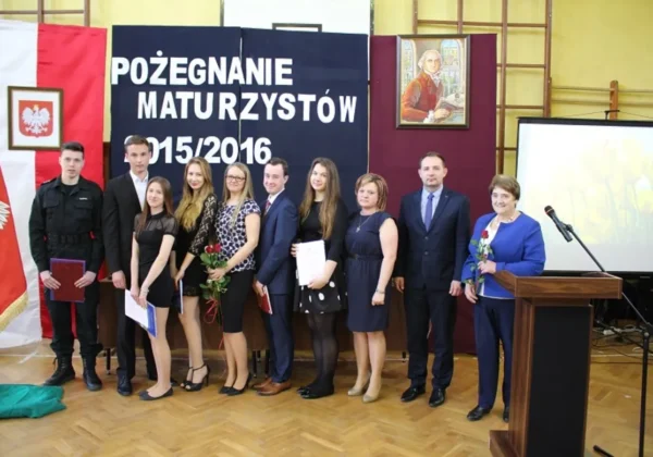 Grupa maturzystów i nauczycieli stoi na scenie podczas uroczystości Pożegnanie Maturzystów 2015/2016, trzymając dyplomy i kwiaty, na tle flagi i prezentacji.