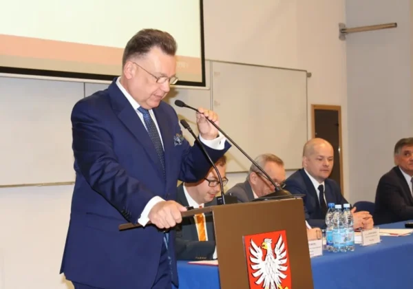 Mężczyzna w granatowym garniturze wygłasza przemówienie przy mównicy z godłem Polski, podczas konferencji z siedzącymi panelistami w tle.