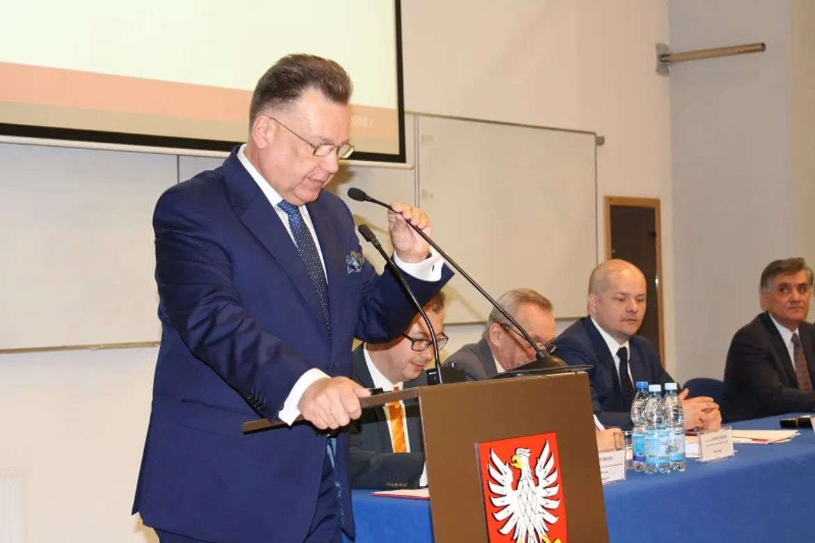 Mężczyzna w granatowym garniturze wygłasza przemówienie przy mównicy z godłem Polski, podczas konferencji z siedzącymi panelistami w tle.
