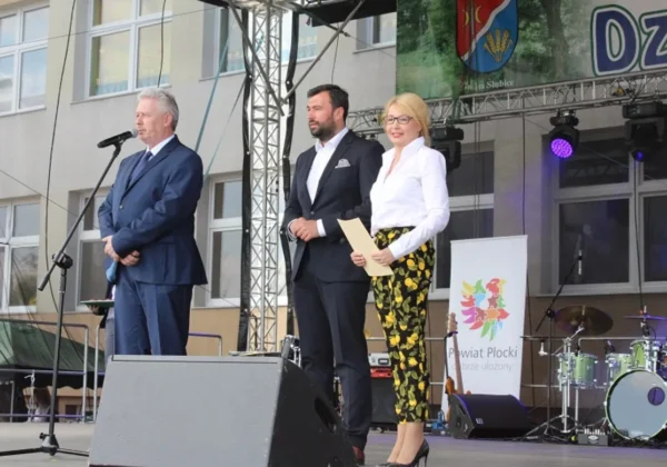 Trzy osoby stoją na scenie podczas XVII Powiatowego Dnia Ziemi w Słubicach; mężczyzna z mikrofonem po lewej, dwóch prezentujących w garniturach po środku, a w tle baner Powiat Płocki i zestaw perkusyjny.