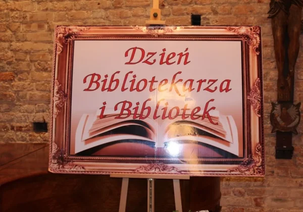 Plakat z napisem „Dzień Bibliotekarza i Bibliotek” z otwartą książką, umieszczony na sztaludze przed ceglaną ścianą.