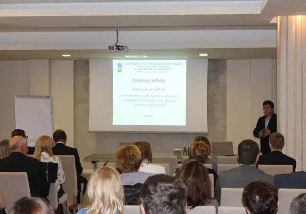Sala konferencyjna z prelegentem stojącym po prawej stronie obok ekranu z prezentacją dotyczącą termomodernizacji budynków użyteczności publicznej; w sali siedzi grupa słuchaczy.