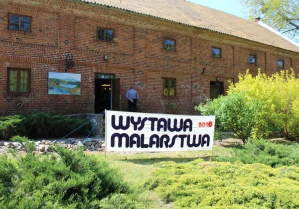 Ceglany budynek z kilkoma oknami, przed którym wywieszono banner WYSTAWA MALARSTWA 2016 zapowiadający plenerową wystawę, z obrazem na ścianie i zielonymi krzewami w ogrodzie.