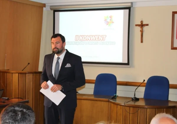 Mężczyzna w eleganckim garniturze stoi z kartkami w dłoniach podczas II Konwentu Samorządów Powiatu Płockiego, w tle wyświetla się prezentacja na projektorze w sali konferencyjnej.