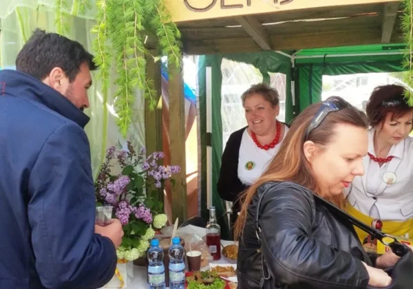 Stoiska podczas XXV Giełdy Rolniczej w Łącku: mężczyzna w granatowej kurtce i kobieta z torebką stoją przy stoliku z kwiatami, napojami i przekąskami, obsługiwane przez dwie kobiety w białych bluzkach z czerwonymi koralami.