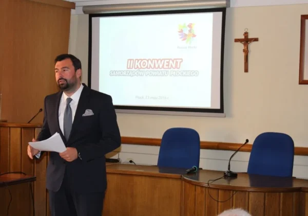 Mężczyzna w garniturze przemawia do uczestników podczas II Konwentu Samorządów Powiatu Płockiego, stojąc przy mównicy z dokumentami, a w tle widać projektor z napisem konwentu i krzyż na ścianie.