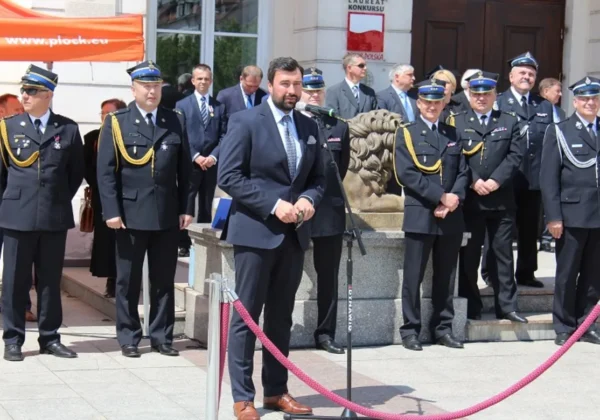 Mężczyzna w garniturze przemawia do mikrofonu podczas Dnia Strażaka 2016, stojąc przed grupą strażaków w mundurach.