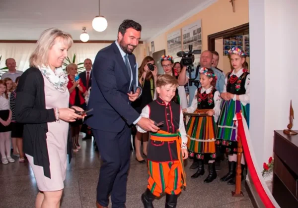 Chłopiec w barwnym ludowym stroju przechadza się po sali podczas VII Zjazdu Stowarzyszenia Rodzina Szkół Chopinowskich w Kobylnikach, otoczony przez dorosłych w garniturach i osoby w tradycyjnych strojach.