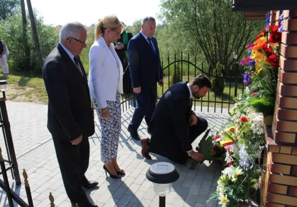 Grupa osób w eleganckich strojach składa kwiaty przy ścianie ozdobionej kwiatami i wieńcami, podczas ceremonii na zewnątrz związanej z oceną stanu bezpieczeństwa powodziowego w powiecie płockim.